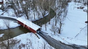 Red covered bridge in winter snow with cars. Beautiful winter scene with river and bare woods and forest trees. Aerial view.