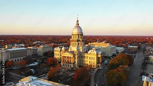 Stunning establishing aerial drone shot of the Illinois State Capitol Building in Springfield, Illinois, at dawn in November as the sun rises and fall leaves glow orange in the distance.