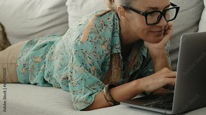 Woman and computer in indoor leisure activity wireless connection. One young adult female people use laptop laying on her stomach on the sofa at home in living room. Lady wearing glasses and writing