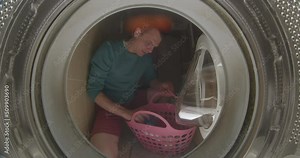 A male cleaner throws dirty laundry into the washing machine. He throws clothes with his hand, closes the glass cover. Start washing and cleaning.