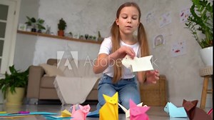 Remote origami lessons. Child smiles show what you need for paper animals and record it on smartphone camera. Little girl shooting online vlog at home. Internet video tutorial or blogging.