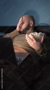 Young bearded man using laptop at night, watching tv at home. Guy watching scary movie on a computer. Shocked, surprised and screaming