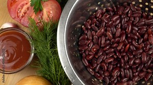 Soaked red beans in a colander, a bunch of parsley and dill, tomato, tomato paste, an onion and garlic lie on a wooden table. Ingredients for preparing a bean dish.