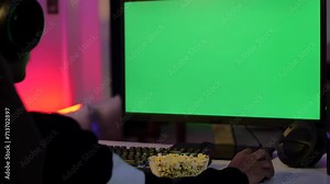 A young gamer watching the green screen - computer monitor with green chroma watching movie. Over-the-shoulder shot of a man with chroma screen while eating popcorn - young man wearing headset pl...
