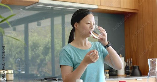 Asian woman standing at kitchen counter after inspecting pill, holding glass, swallowing water
