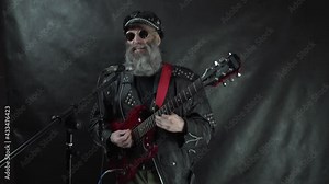 Rockstar bearded in leather jacket and cap wearing round sunglasses plays red electric guitar and sings on stage with black background
