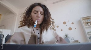 Low angle view of curly-haired young woman covered with plaid sitting on sofa at home, checking her temperature using thermometer while having online talk to doctor using PC