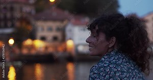Curly haired woman tourist smiles observing a traditional village in the Basque Country illuminated with lights at night