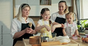 Happy daughters with braids sisters helping granny taking flour from back pouring on sieve sifting through. Middle aged granny teaching offsprings baking cooking homemade tasty cookies pasta pizza.