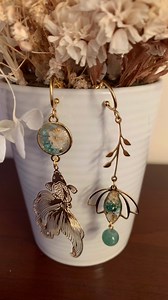 🐠stunning 24K gold plated hoops one with a goldfish hanging of a pretty golden connector with teal and white flowers and hints of gold foil and the other with matching bead on a lotus flower with matching flowers 🪷 | Sophie’s creative studio