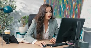 Focused on work attractive girl in classic makeup wearing a shirt and jacket, sits at a desk in a company in front of a computer, fills out documents, reads email, answers messages, smiling at camera