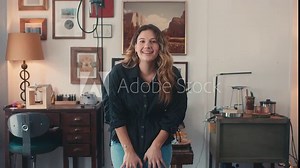 Woman sitting in rustic studio with art on walls looks up and smiles, Zoom Out