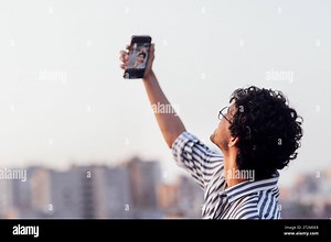 Young curly-haired guy in glasses and striped shirt answers video call on phone. Smiling mixed-race man shows victory symbol and takes selfie standing Stock Photo - Alamy