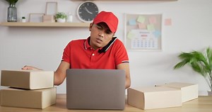 Download Portrait of Young Asian busy postman in red uniform and cap sitting at desk and talking on mobile phone in postal office store and working at laptop. Parcels front side. for free