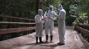 Three men in biohazard suits and masks standing on a bridge over a stream preparing to sample the water for contaminants
