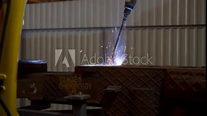 Industrial robotic arm performs precision arc welding on metal components, guided by skilled operator in manufacturing plant. Automated machinery ensures accurate joinery, sparks fly during process.