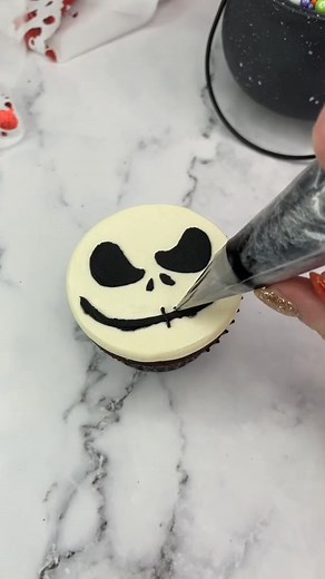 Jack Skellington cupcakes 🖤 Which character should I make next? 🤍 Use a large round piping tip (ateco 809) to add white buttercream onto the cupcakes. 🖤 Press the buttercream down onto parchment paper to achieve a flat surface. 🤍 Freeze the cupcake for 15 minutes. 🖤 Remove the parchment paper and attach your stencil to the top. I made the stencil using a square of parchment paper and a sharp blade. 🤍 Apply a thin layer of black buttercream over the stencil. 🖤Then carefully peel it off. Ad