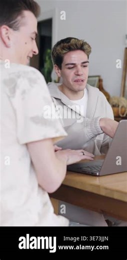 Two cheerful young men using a laptop together while sitting at a wooden table. They are discussing and pointing at the screen at home. Vertical footage Stock Video Footage - Alamy