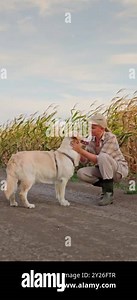 A woman farmer enjoys a playful moment with her dog beside a cornfield, embodying the sweet and simple pleasures of rural life. The golden cornfield sways gently in the background, creating a serene Stock Video Footage - Alamy