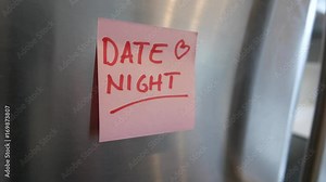 Putting a Date night sticky note reminder on a fridge. Closeup on the hand and paper.