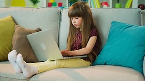 Cute little girl thinking about something and writing down it in laptop computer. Little girl has an idea. Sitting on the couch