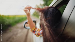 Rear view female passenger sitting put head out of window. Close-up brunette loose long healthy hair fluttering in wind.Young woman enjoying ride, touching hand her hairstyle. Driving at speed in car.