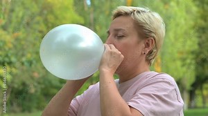 A blonde girl with short hair inflates a balloon against the background of green nature close-up