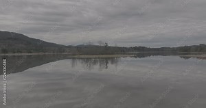 Ungraded 4k low altitude wide-angle drone footage of a miniature island with deciduous trees in the middle of a reflective lake on a cloudy morning at Alder Lake in Washington State.