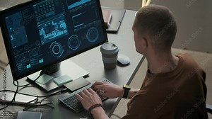 High angle back view shot of young male Caucasian programmer sitting at desk in front of PC and working with complex data in modern office at nighttime