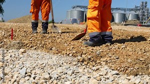 Low angle view on two workers using shovels to set up level foundation to right measures leveling layer of fraction, small crushed stone at building site.
