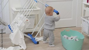 Cute little toddler helping mother with household chores, child playing with clothes drying rack in living room, kid having fun exploring all around the house, 9 month old caucasian baby boy at home.