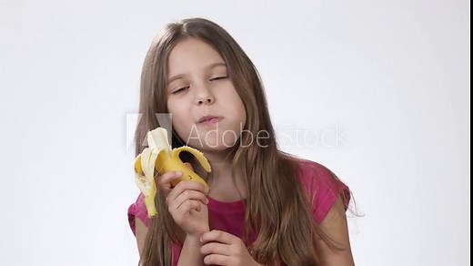 Little girl eating a ripe yellow banana on a white background. The girl bites a banana and tastes it.