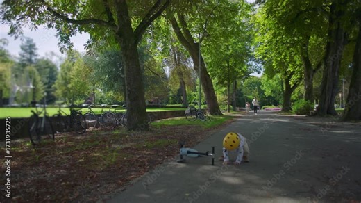 Toddler falling off scooter on park path while wearing helmet, child losing balance during playtime accident in outdoor environment