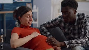 Close up of interracial couple with pregnancy at home holding hands on belly. Mixed race young partners expecting child and touching baby bump while sitting on living room couch