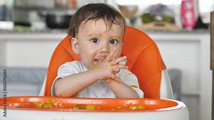 Funny kid sits in a chair for feeding children and claps his hands, eats himself, got dirty all over the food. The child learns to eat on his own. Healthy food for preschool children. Smart kid.