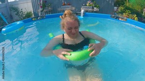 A man's hand splashes in the face of a plump woman who is swimming in the pool sitting in a swimming noodle. The woman smiles and squirts her face. First-person view