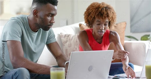 African American couple working, woman flipping papers urging man to type and check laptops on sofa