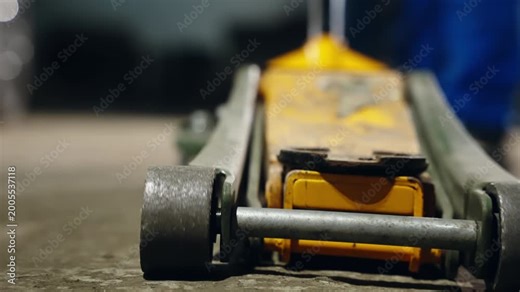 Close-up and operational view of a yellow hydraulic floor jack used by a mechanic on a concrete workshop surface to lift a vehicle, showing metal construction and handle mechanism.