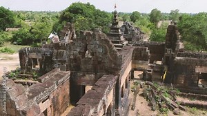 Aerial view flying low over ancient ruined Angkor temple (Buddhist wat) in Cambodia, Southeast Asia; large blocks, colonnades and Hindu style peak are visible.