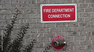 fire department connection sign on beige brick wall with two twin connections below from one spout in red circle auto spkr, white on red Stock Video
