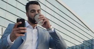 Bearded Handsome Man Wearing light Formal Suit Standing in front of Camera. Using Mobile Phone and reading Messages on good mood. Good looking Businessman drinking hot Coffee from paper cup