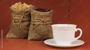 Coffee is poured into a cup and behind stands coffee beans in burlap bag and next sugar in burlap bag on brown background