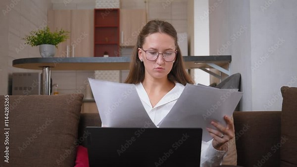 Businesswoman sitting on sofa in home office, looking at documents in front of laptop with serious expression. Focused and engaged, she analyzes paperwork, coping with deadlines and stress