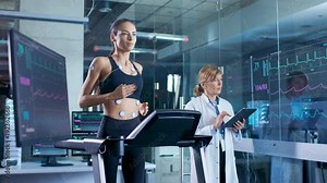 Beautiful Woman Athlete Runs on a Treadmill with Electrodes Attached to Her Body, Female Physician Uses Tablet Computer and Controls EKG Data Showing on Laboratory Monitors.