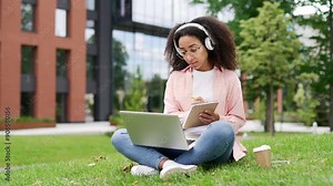 African american female student listens to online video course using laptop sitting on lawn in campus near university building. Young black woman in headphones at remote learning training taking notes