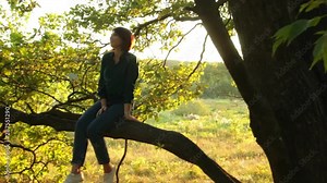 Girl sitting and swinging on a branch of a tree. Young woman in a forest having fun.
