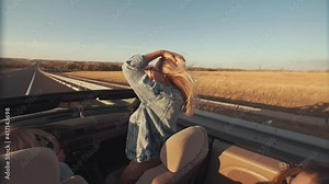 View from back seat as young women enjoy lifestyle during ride in convertible, women in sunglasses raise their hands, have fun and fool around. Ride in convertible on highway, sunset