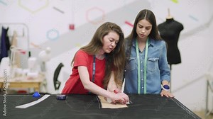 Tracking shot of professional clothing designer teaching young woman how to draw chalk draft on fabric using paper pattern and sewing curve ruler
