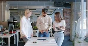 Working team of man and two women standing over the table with tablet device and charts and discussing project. IT employees. Mixed-races male and females talking and brainstorming. Startup concept.