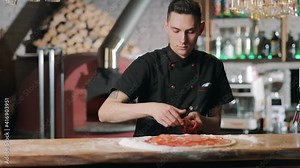 Male chef in black tunic putting red sweet pepper on pizza base. Skillful pizzaiolo adding raw ingredients on dough. Concept of culinary.